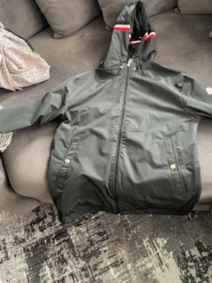 Svart vindjacka från Moncler nfc funkar - Svart vindjacka med dragkedja, huva och röda samt vita ränder på huvan. Två framfickor med tryckknappar och diskret logga på ärmen. Jackan har en sportig look och är tillverkad i ett vattenavvisande material.