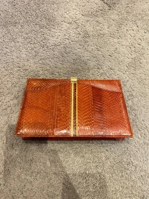 Brun kuvertväska i ormskinnslook - Snygg brun kuvertväska med ormskinnsmönster och guldfärgad detalj på locket. Väskan har en rektangulär form och stängs med ett metallspänne. Perfekt accessoar för dig som vill sticka ut med en unik och trendig look.