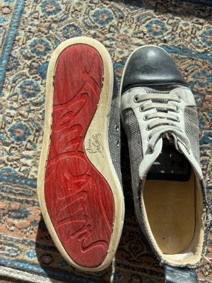 Christian Louboutin svarta sneakers - Snygga sneakers från Christian Louboutin med klassisk röd sula, svart tåparti och häl, samt gråmelerad textil på sidorna. Skorna har vita snören och detaljer i läder och textil. Perfekta för dig som vill sticka ut med exklusiv design.