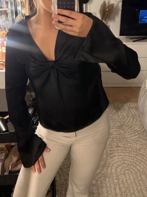Svart satinblus med v-ringning - Stilren svart blus i glansigt satinmaterial med djup v-ringning och twistad detalj framtill. Blusen har långa, vida ärmar och en avslappnad passform som ger en trendig look. Perfekt att matcha med både jeans och. 