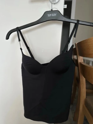 Svart linnetopp med strassdetaljer - Snygg svart linnetopp med tunna axelband och strassdetaljer på banden. Toppen har en figurnära passform och lätt vadderade kupor för extra stöd. Perfekt att styla med jeans eller kjol för en trendig look.