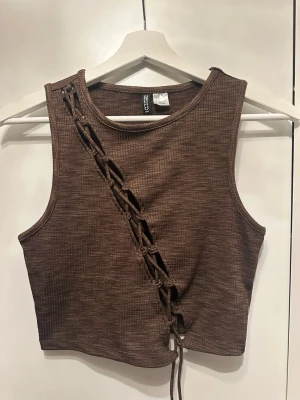 Brun ribbad linnetopp med snörning - Trendig brun croppad linnetopp i ribbad bomullsblandning. Toppen har en unik snörningsdetalj diagonalt framtill och är ärmlös med rund halsringning. Perfekt för att lyfta din outfit med en edgy touch.