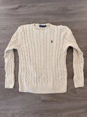 Kabelstickad beige tröja Polo Ralph Lauren - Kabelstickad beige tröja från Polo Ralph Lauren med rund halsringning och den klassiska broderade loggan på bröstet. Tröjan har långa ärmar och ribbade muddar vid ärmslut och nederkant. Perfekt för dig som gillar tidlös och clean stil.