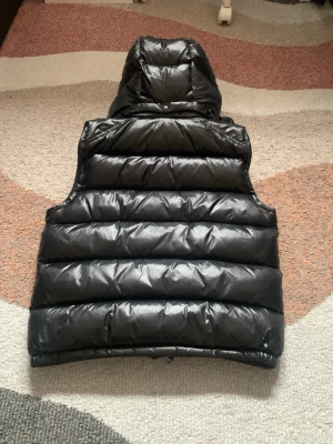 Svart dunväst med huva - Snygg svart dunväst med glansig finish och stor vadderad huva. Västen har breda quiltade paneler och dragkedja framtill. Perfekt för lager-på-lager och ger en cool streetwear-look. Passar dig som vill ha både stil och värme under kyliga dagar.