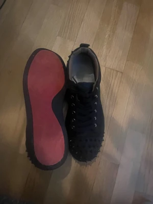 Svarta sneakers med nitar och röd sula Louboutin  - Säljer ett par svarta sneakers med coola nitar på tån och sidorna. Skorna har en klassisk låg profil och en ikonisk röd sula. Svarta skosnören och metallhål för snörningen. Perfekta för dig som vill sticka ut lite extra. OBS andvända endast 5 gånger.