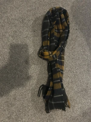 Rutig halsduk från Barbour - Säljer en snygg rutig halsduk från Barbour i lammull och kashmir. Färgerna är svart, grå, senapsgul och vit med klassiskt rutmönster och fransar i ändarna. Perfekt accessoar för att lyfta din outfit.