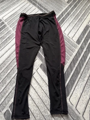 Svarta leggings med vinröda detaljer - Säljer ett par svarta leggings med vinröda paneler längs sidorna och meshdetaljer vid bensluten. De har en sportig look och är tillverkade i stretchigt material som passar perfekt för träning eller chill. Snygga kontrastsömmar längs benen.