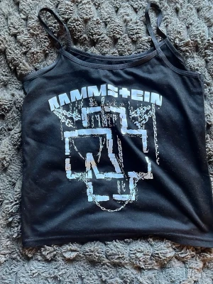 Svart Rammstein linne - Säljer en svart linne med Rammstein-tryck framtill. Toppen har tunna axelband och ett stort vitt tryck med bandets logga och kedjedetaljer. Perfekt för dig som gillar bandmerch och vill ha en edgy stil.