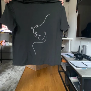 Svart t-shirt med konstnärligt tryck - Svart t-shirt med ett stilrent, konstnärligt tryck i vitt som föreställer ett ansikte i linjeart. T-shirten har korta ärmar och en klassisk rund hals. Perfekt för dig som gillar minimalistisk design och vill sticka ut med en unik look.