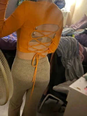Orange croppad topp med snörning - Trendig orange croppad topp med lång ärm och ribbad struktur. Baksidan har en öppen design med snygg snörning som knyts i midjan. Perfekt för dig som vill sticka ut med en cool detalj på ryggen.