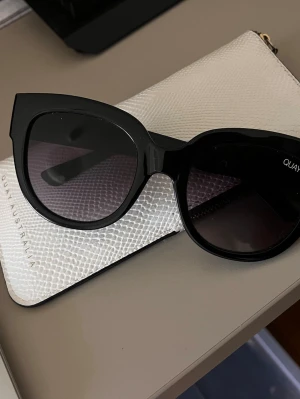 Svarta cat eye-solglasögon Quay - Stilrena svarta solglasögon från Quay med trendig cat eye-form. Bågarna är i blank svart plast och glasen är mörka, vilket ger en cool och modern look. Perfekta för att lyfta vilken outfit som helst.