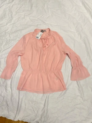 Ljusrosa blus med volangärm från H&M - Supersöt ljusrosa blus från H&M med långa volangärmar och knytning vid halsen. Blusen har resår i midjan för en snygg passform och är gjord i ett lätt, luftigt tyg som känns skönt mot huden. Perfekt för en trendig och feminin look.