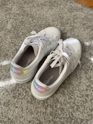 Vita Adidas sneakers med holografiska detaljer - Snygga vita sneakers från Adidas med klassiska tre ränder i holografiska färger på sidan och matchande holografisk detalj på hälen. De är i nyskick förutom ett litet märke så ena hälen som man kan se på sista bilden!