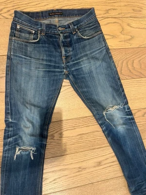 Blå slitna jeans från Nudie Jeans - Säljer ett par blå jeans från Nudie Jeans med skinny fit och slitningar på båda knäna. Jeansen har klassisk femficksdesign, orangea sömmar och är tillverkade i denim. Perfekta för en avslappnad och trendig look.