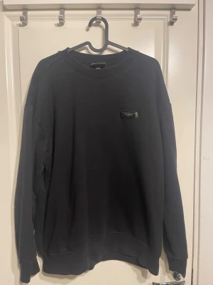 Svart sweatshirt från HM - Svart sweatshirt från HM med rund halsringning och diskret logotyp på bröstet. Tröjan har en klassisk passform med ribbade muddar vid ärmslut och nederkant. Perfekt för en avslappnad och stilren look.
