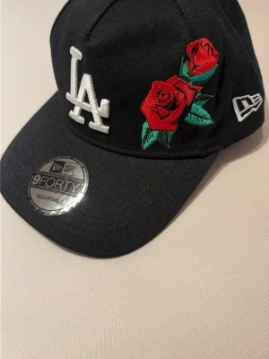 Svart LA keps med rosbroderi - Cool svart keps från New Era med LA-logga i vitt och två röda rosor broderade på framsidan. Modellen är 9FORTY och har justerbar passform. Materialet är bomull och kepsen har böjd skärm samt broderad New Era-logga på sidan.