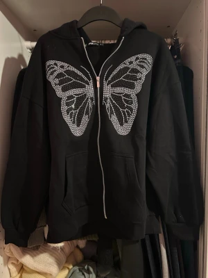 Svart hoodie med fjäril från FB Sister - Säljer en svart hoodie med dragkedja från FB Sister. På framsidan finns en stor fjäril i silvriga nitar som ger en cool vibe. Hoodien har huva och fickor framtill. Perfekt för dig som gillar streetstyle och vill sticka ut lite extra. Stor i storleken