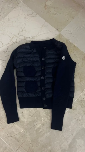Mörkblå cardiganjacka från Moncler - OBS! (Kan senast skicka den 18 Juni, pågrund av resa) Det Snygg svart cardiganjacka från Moncler med quiltad front och stickade ärmar. Jackan har knappstängning, ribbade muddar och Moncler-logga på ärmen. Materialmix av dun och ull ger både värme och stil. Perfekt för lager på lager och streetwear-vibe.