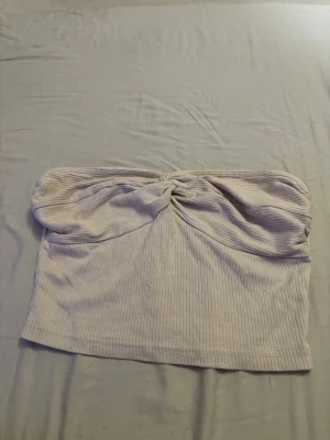 Ljusbeige ribbad tubtopp med twist - Trendig ljusbeige tubtopp i ribbat material med snygg twistdetalj framtill. Toppen är axelbandslös och har en croppad passform som passar perfekt till höga jeans eller kjol. Säljer denna pågrund av av den inte kommer till användning 