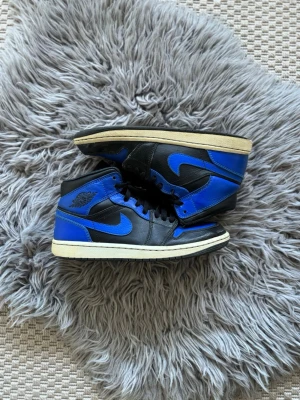 Air Jordan 1 Retro High OG Royal - Snygga Air Jordan 1 Retro High OG i svart och blått läder med klassisk vit sula och svarta skosnören. Ikonisk vinge-logga på sidan och blå Nike swoosh. Perfekta för dig som vill sticka ut med en tidlös sneakerklassiker.