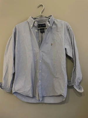 Ljusblå skjorta från Ralph Lauren - Klassisk ljusblå skjorta från Ralph Lauren med button-down krage och broderad logga på bröstet. Skjortan har lång ärm, knappar framtill och är tillverkad i bomull. Perfekt för en clean och stilren look.
