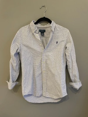 Rutig skjorta från Ralph Lauren - Klassisk vit skjorta från Ralph Lauren med blått rutmönster och broderad logga på bröstet. Skjortan har lång ärm, knappar framtill och en tidlös krage. Perfekt för dig som gillar stilrena och preppy plagg.