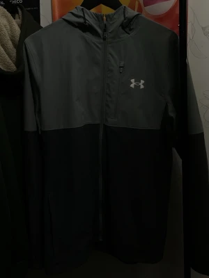 Grå och svart vindjacka Under Armour - Snygg tvåfärgad vindjacka från Under Armour i grått och svart. Jackan har huva, hel dragkedja framtill, bröstficka med dragkedja och Under Armour-logga på bröstet. Perfekt för blåsiga dagar och sportiga outfits. Riktigt bra skick använd kanske tre gånger 