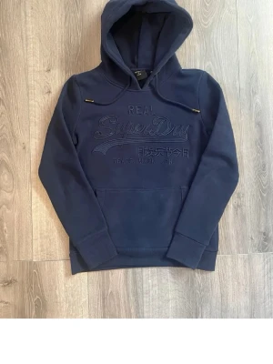 Mörkblå hoodie från Superdry - Snygg mörkblå hoodie från Superdry med broderad logga och text på bröstet. Klassisk känguruficka framtill och justerbar huva med snören. Tillverkad i mjukt material som känns skönt mot huden. Perfekt för en avslappnad och trendig look.