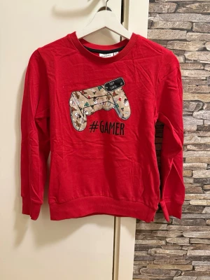 Röd långärmad gamertröja - Cool röd långärmad tröja med ett stort gamer-tryck framtill i form av en spelkontroll och texten #GAMER. Tröjan har rund halsringning och ribbade muddar vid ärmslut och nederkant. Perfekt för dig som älskar gaming och vill sticka ut.