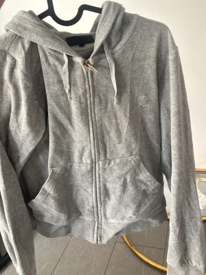 Grå zip hoodie från Juicy Couture - Snygg grå hoodie från Juicy Couture med dragkedja framtill, huva med dragsko och två stora fickor. Tillverkad i mjukt material som känns skönt mot huden. Perfekt för en avslappnad och trendig look.