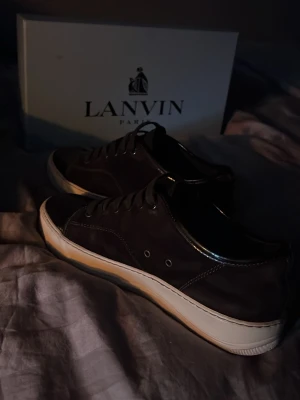 Bruna lanvins med vit sula  - Säljer ett par snygga svarta Lanvin sneakers med vit gummisula och svarta skosnören. Skorna har en låg profil och är tillverkade i läder med diskreta sömdetaljer. Perfekta för dig som vill ha en stilren och exklusiv look. Box kommer med