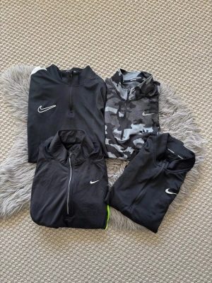 4-Pack Svarta långärmade träningströjor Nike - 4sr Svarta långärmade träningströjor från Nike med halv dragkedja och vit logga på bröstet. Tillverkad i ett lätt och stretchigt funktionsmaterial som passar perfekt för löpning eller gym. Enkel och stilren design med hög krage.