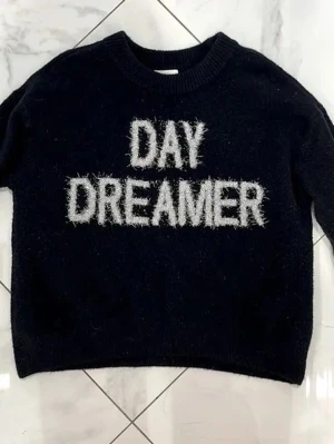 Svart stickad tröja DAY DREAMER - Cool svart stickad tröja med texten 'DAY DREAMER' i vit fluffig stil på bröstet. Tröjan har rund halsringning och långa ärmar. Perfekt för dig som gillar statement-plagg och vill ha en mysig men trendig look.