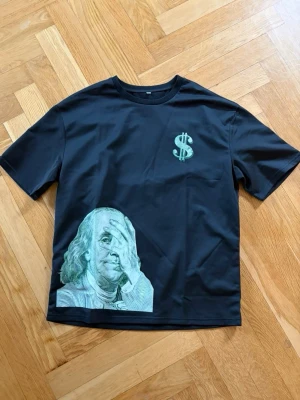 Svart t-shirt med dollartryck - Cool svart t-shirt med ett grönt dollartecken på bröstet och ett stort tryck av Benjamin Franklin i grönt och vitt nedtill. T-shirten har normal passform och korta ärmar. Perfekt för dig som gillar streetwear och statement-plagg.