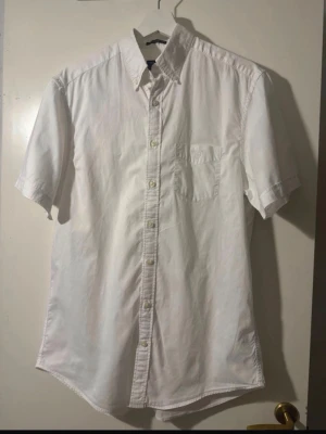 Vit kortärmad skjorta från Gant - Klassisk vit skjorta från Gant med korta ärmar och bröstficka. Skjortan har knappar framtill och broderad Gant-logga på fickan. Perfekt för en clean och stilren look.