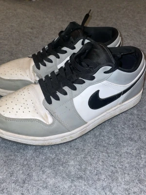 Nike Air Jordan 1 Low grå/vit/svart - Snygga Nike Air Jordan 1 Low sneakers i grått, vitt och svart med klassisk svart swoosh och röd Jumpman-logga på plösen. Skorna har svart snörning och är tillverkade i läder och textil. Perfekta för dig som gillar streetwear och ikoniska sneakers.