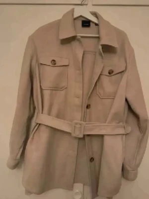 Beige overshirt med bälte storlek 38/40 - Stilren beige overshirt med bälte i midjan och två stora bröstfickor med knappar. Klassisk krage och knäppning framtill. Perfekt lager-på-lager-plagg i mjukt material som ger en avslappnad men trendig look.