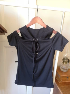 Topp med cutouts och snörning - Svart ribbad topp med cool snörning framtill och cutouts vid axlarna. Kortärmad modell med en snygg, sexig, lite edgy look. Perfekt för dig som vill sticka ut med detaljer och gillar trendiga toppar. Köpt i Polen. Säljes pga garderobs rensning. 