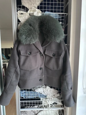 Grå kort kappa med pälskrage Meotine - Snygg grå kappa från Meotine i kort modell med stora bröstfickor och knappar framtill. Kappan har en lyxig, bred pälskrage (äkta päls) De går inte och sätta fast med dom knapparna jag har ringat in men de är absolut inget som påverkar pälsen. 3000kr nypris 