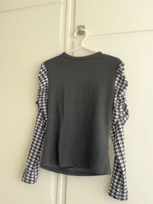 Grå topp med hundtandsmönstrade ärmar - Cool grå långärmad topp med svartvita hundtandsmönstrade ärmar. Perfekt för dig som vill sticka ut med en unik och trendig look. Rak passform och rund halsringning. Passar till både jeans och kjol.