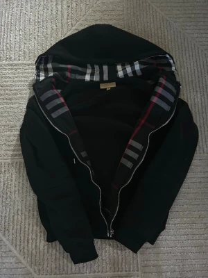 Svart hoodie från burberry med dragkedja - Snygg svart hoodie från burberry med dragkedja och klassiskt rutigt foder i huvan. Diskret burberry-logga på bröstet och ribbade muddar vid ärmar och nederkant. Perfekt för en chill och stilren look. Väldigt skön tyg.