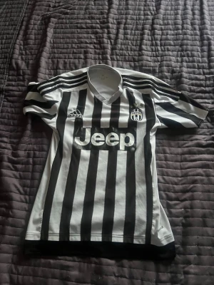 Juventus matchtröja svartvit Adidas - Juventus officiella matchtröja från Adidas i klassiskt svartvitt randigt mönster. Tröjan har klubbmärke, Adidas-logga och Jeep som sponsortryck på bröstet. Korta ärmar och V-ringad krage. Perfekt för fotbollsfans!