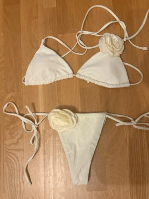 Vit bikini med blommor - Supersöt vit bikini med stora dekorativa blommor på både överdel och underdel. Klassisk trekantsmodell med tunna band som knyts i nacken och ryggen. Perfekt för sommaren! Skriv vid eventuella frågor💞