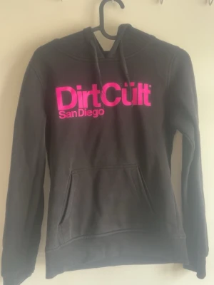 Svart Dirt Cult hoodie med rosa tryck - Svart hoodie från Dirt Cult med stor magficka och huva. Framtill finns ett fett rosa tryck med texten 'Dirt Cult San Diego'. Hoodien har dragsko i huvan och är gjord i mjukt material, perfekt för chill dagar.