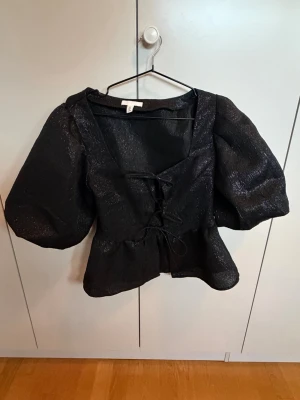 Svart glittrig blus med puffärmar - Säljer en svart blus från H&M med glittrigt tyg och puffiga ärmar. Blusen har en snygg snörning framtill och markerad midja som ger en cool siluett. Perfekt för dig som gillar trendiga detaljer och att sticka ut.