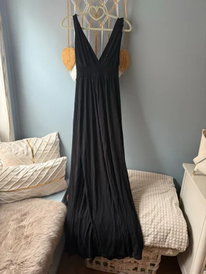 Svart långklänning med v-ringning - Elegant svart långklänning med djup v-ringning och breda axelband. Klänningen har ett flödande fall och är tillverkad i ett mjukt material som ger en snygg siluett. Perfekt för dig som vill ha en stilren och klassisk look Passar både S-M.