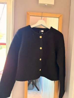 Svart kofta med guldknappar - Snygg svart kofta med rund halsringning och fyra guldiga knappar framtill. Koftan har en rak, något kortare passform och långa ärmar. Perfekt att slänga över en t-shirt eller klänning för en stilren look. Kan användas som en jacka under vår /sommar. Aldrig använt! 