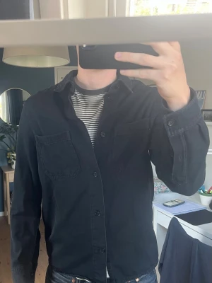 Svart overshirt med fickor från arket  - Stilren svart overshirt med två stora bröstfickor och knäppning framtill. Skjortan har klassisk krage och raka ärmar. Perfekt att bära över en t-shirt för en avslappnad look. 