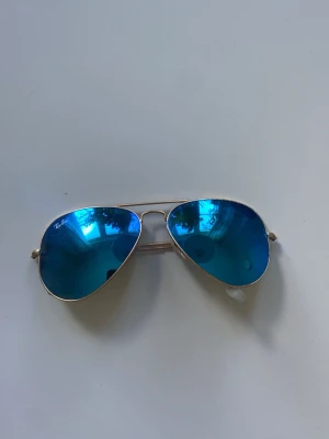Ray-Ban aviator solglasögon blå - Snygga aviator solglasögon från Ray-Ban med spegelblå glas och tunn guldfärgad metallbåge. Klassisk pilotmodell som ger en cool vibe och passar perfekt till sommaren. Ray-Ban-loggan syns på linsen. Lätta och bekväma att bära.