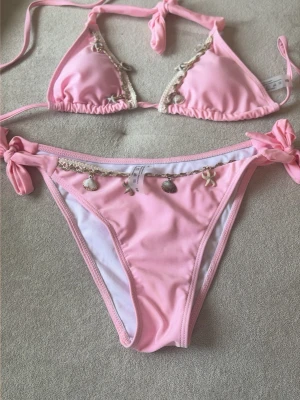 Rosa bikini med snäckdetaljer - Supersöt ljusrosa bikini med snäckformade berlocker och beige spetsdetaljer. Både överdel och underdel har knytband för justerbar passform. Perfekt för sommaren och strandhäng!